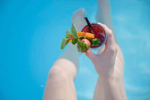 Hands holding tropical cocktail with fresh mint and fruit over blue pool water