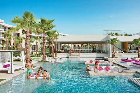 Resort pool with palm trees, modern white architecture, and guests lounging with pink accent pillows