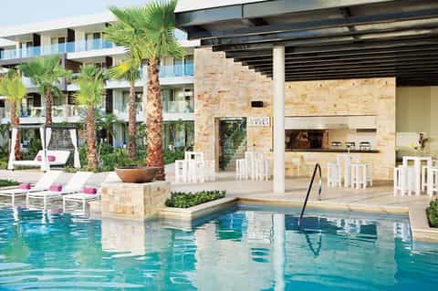 Resort pool deck with modern architecture, palm trees, stone fireplace, and lounge seating