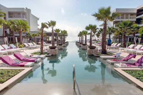 Luxury resort pool with magenta loungers, palm trees, and flowing water channels between modern white buildings