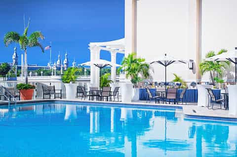 Swimming pool with blue water, white umbrellas, palm trees, and castle-like building in distance