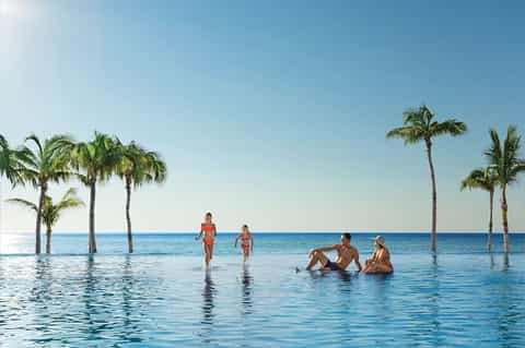 Infinity pool with guests swimming and palm trees overlooking a blue ocean horizon