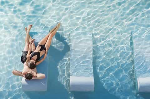 Overhead view of two people floating on white platforms in crystal-clear turquoise pool water
