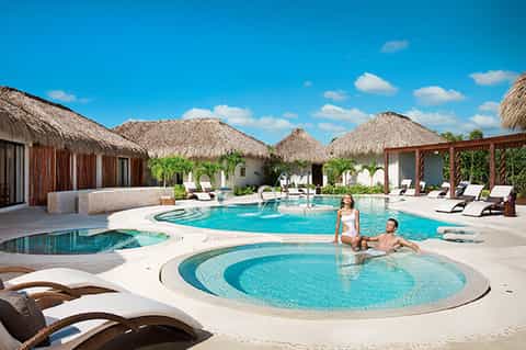 Tropical resort pool with thatched-roof bungalows, jacuzzi, and guests enjoying paradise setting