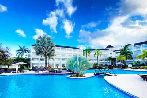 Resort-style pool surrounded by white buildings, palm trees, and colorful lounge chairs under blue sky