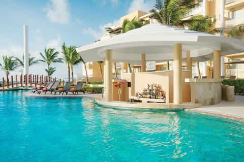 Resort pool bar with turquoise water, white covered bar structure, palm trees, and ocean view