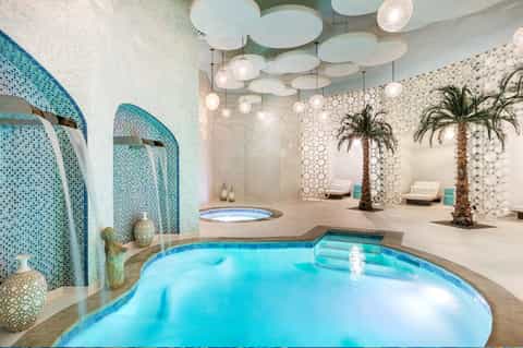 Luxurious indoor spa pool with turquoise water, glass mosaic walls, and palm trees