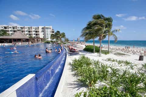 Beachfront infinity pool with thatched-roof bar, white hotel building, and ocean view