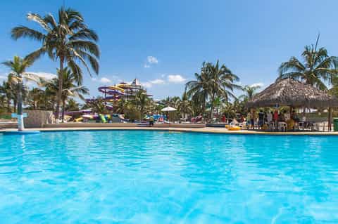 Bright turquoise resort pool with water slides, palm trees, and recreational facilities under clear sky