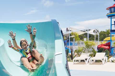 Child sliding down turquoise water slide at outdoor water park with palm trees