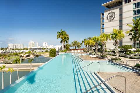 Long narrow resort pool with palm trees, modern white building, and marina view in background