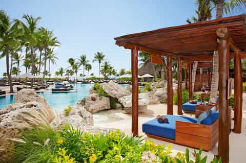 Luxury beach resort with wooden cabana, blue cushions, lagoon pool, palm trees, and umbrellas