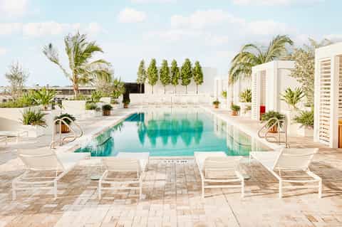 Modern resort pool with white lounge chairs, tropical landscaping, and buildings