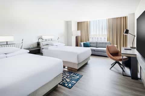 Modern hotel room with two beds, gray seating area, large windows with curtains, desk, and contemporary furnishings