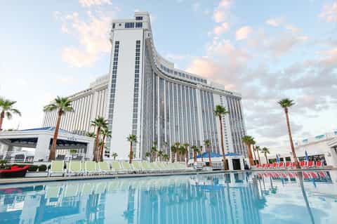 Tall curved white hotel building with palm trees reflected in resort pool and lounge chairs