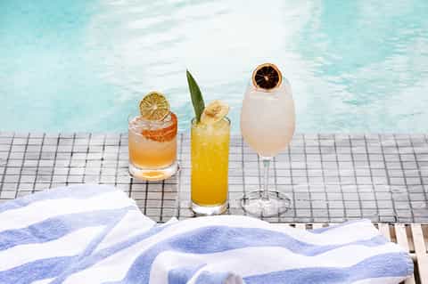 Three colorful cocktails with citrus garnishes on pool deck overlooking turquoise water