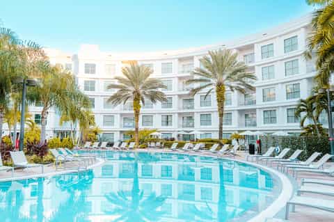 Curved resort pool with white lounge chairs, palm trees, and modern white building