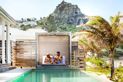 Couple relaxing in poolside cabana overlooking turquoise pool with mountain backdrop and palm trees