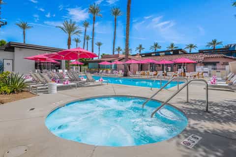 Resort pool complex with hot tub, pink umbrellas, and palm trees under blue sky