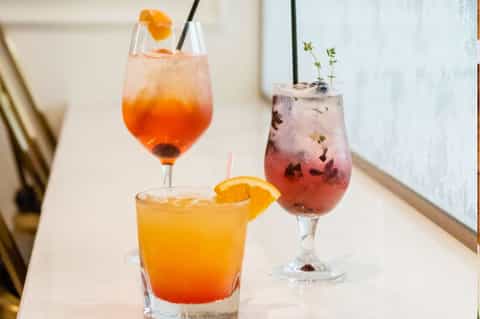 Three colorful cocktails with orange and red hues, garnished with citrus slices and fresh herbs