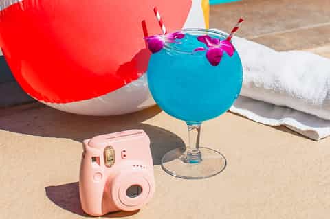 Bright blue tropical cocktail with pink orchid flowers on sandy beach next to pink camera and red beach bag