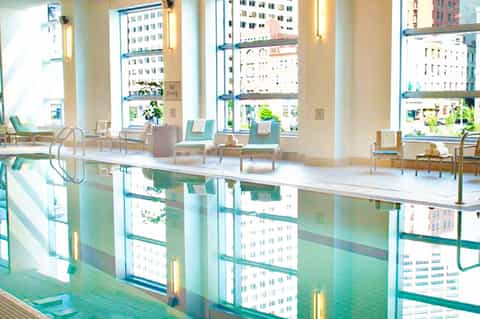 Indoor lap pool with floor-to-ceiling windows overlooking a city skyline, with lounge chairs nearby