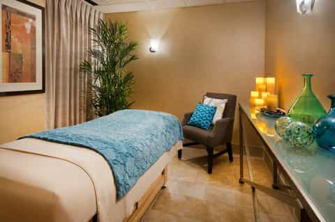 Warm-toned bedroom with teal quilted bedding, accent chair, and decorative glass vases on nightstand.