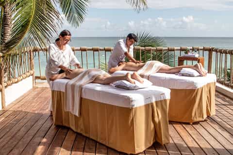 Beachfront spa treatment with two massage therapists working on clients overlooking ocean with palm fronds