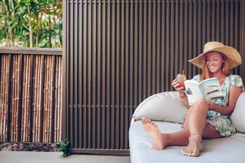 Woman relaxing on white lounge chair reading book, wearing straw hat near black fence