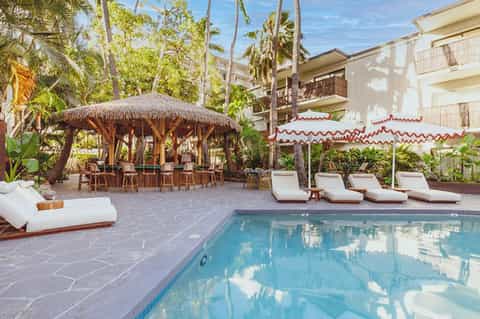 Resort pool deck with white lounge chairs, thatched roof structure, palm trees, and umbrellas