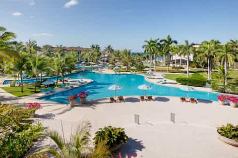 Large resort pool complex with multiple sections, palm trees, and beachfront hotel buildings