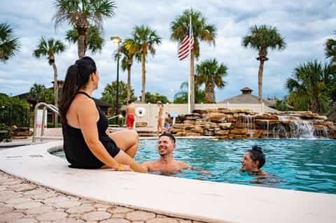 Family enjoying resort pool with waterfall feature, palm trees, and American flag in background
