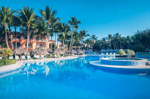 Large resort pool complex with palm trees, thatched umbrellas, Spanish colonial buildings, and clear sky