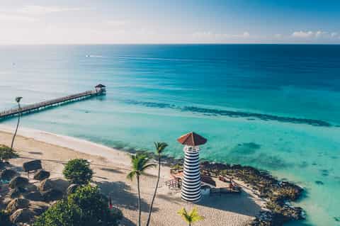 Tropical beach with turquoise water, wooden pier, striped lighthouse, and palm trees