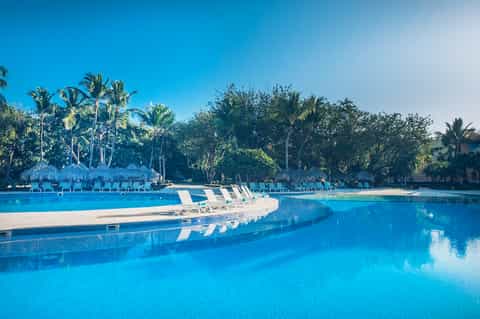 Resort lagoon pool with white lounge chairs, tiki huts, palm trees, and turquoise water