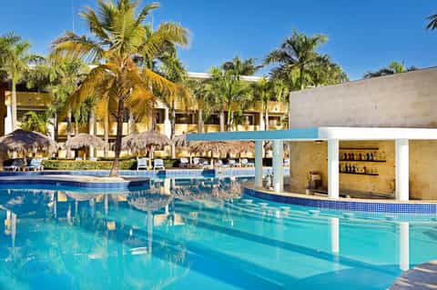 Resort pool with thatched umbrellas, palm trees, lounge chairs, and swim-up bar structure