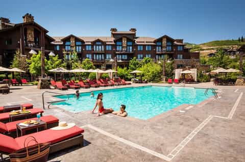Resort pool with loungers and umbrellas, guests swimming, large lodge building with stone accents on hillside
