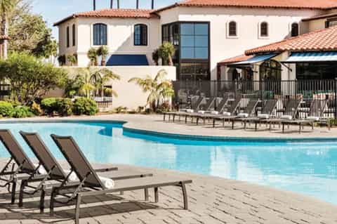 Spanish-style resort pool with lounge chairs, white building, palm trees, and covered patio area