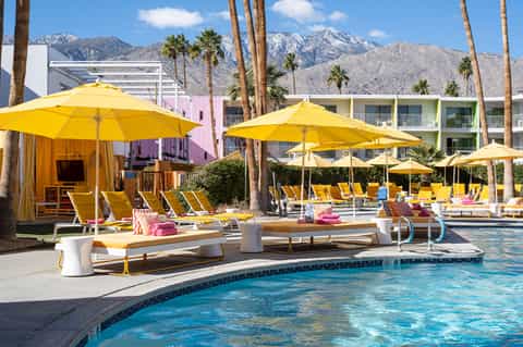Vibrant resort pool with bright yellow umbrellas, loungers, pink accent buildings, and mountain backdrop
