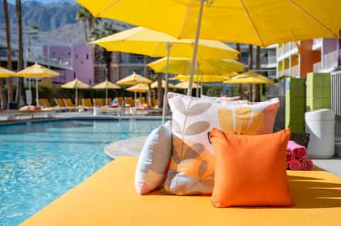 Pool lounge chairs with colorful pillows under yellow umbrellas with mountain backdrop