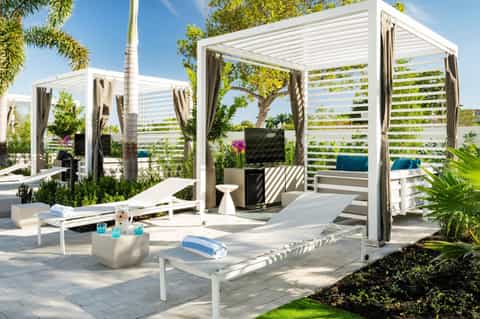 Upscale outdoor lounge with white pergolas, draped curtains, lush landscaping, and contemporary furniture