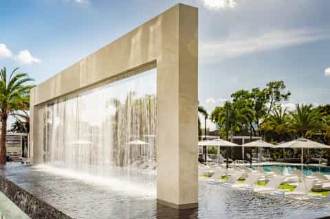 Modern waterfall feature cascading over geometric concrete structure with resort pool and umbrellas in background