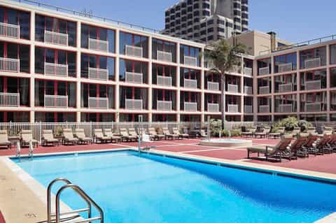 Large rectangular pool with loungers on red deck in front of modern multi-story hotel building