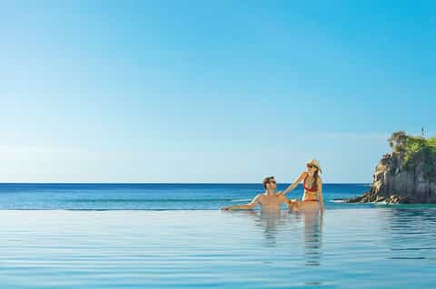 Couple wading in crystal clear shallow water with rocky cliffs and ocean backdrop