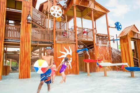 Children playing in shallow water at a wooden water park structure with colorful beach balls and water features