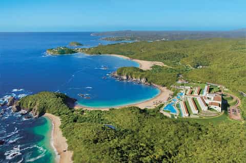 Aerial view of beachfront resort with colorful buildings nestled in green forest along curved coastline
