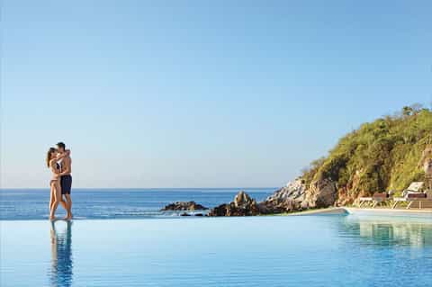 Couple wading in infinity pool overlooking ocean and rocky coastline at golden hour