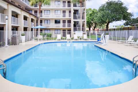 Resort pool surrounded by multi-story buildings with balconies, white lounge chairs, and green landscaping