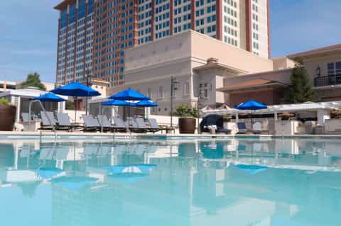 Resort pool with blue umbrellas, lounge chairs, tall building behind, and clear blue sky