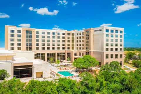 Modern tan hotel with turquoise pool, green landscaping, and blue sky with clouds